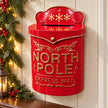 Red metal North Pole Express Mail letterbox with gold lettering and snowflakes, styled on a cream wall with Christmas greenery.