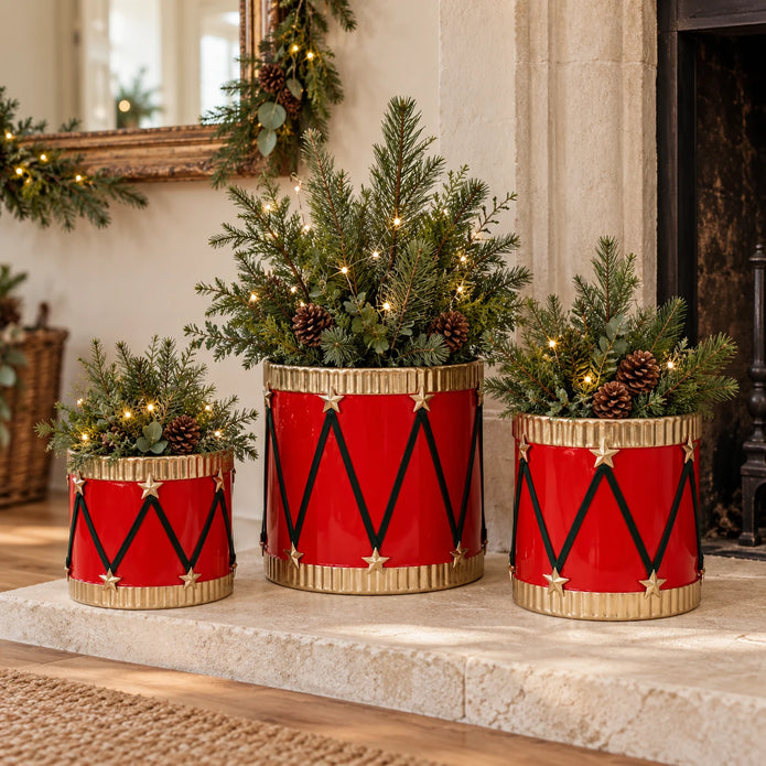 Three red and gold drum planters with winter greenery beside a pale stone fireplace.