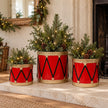 Three red and gold drum planters with winter greenery beside a pale stone fireplace.