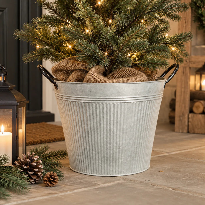 Round ribbed zinc metal planter with black handles holding a small lit fir tree by a country home entrance