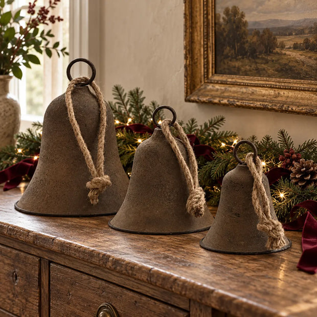 Rustic brass-coloured decorative bells in three sizes styled with evergreen garland and burgundy ribbon on an oak console