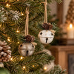 Set of two small rustic hanging Christmas bells, silver grey and whitewashed, with pinecones and greenery on a Christmas tree