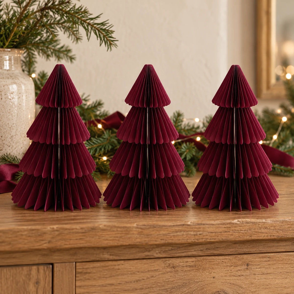 Three matching 20cm burgundy paper Christmas trees on an oak sideboard with festive greenery.