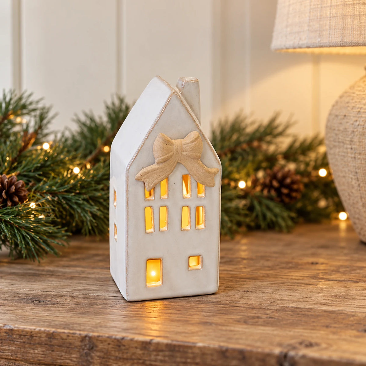 Small ceramic LED house with bow detail glowing on an oak console in a cream Christmas hallway.
