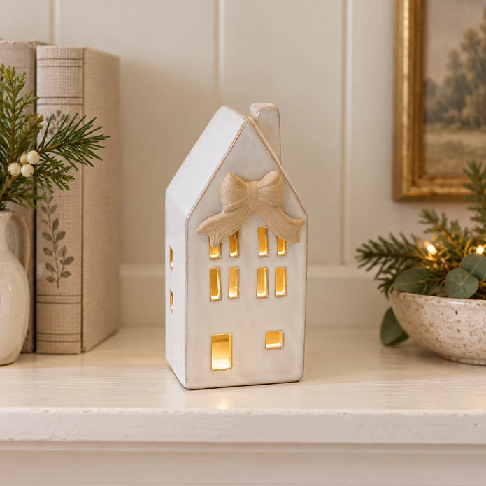 Small light-up ceramic house with its beige bow styled on a cream shelf with linen-bound books.