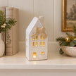 Small light-up ceramic house with its beige bow styled on a cream shelf with linen-bound books.