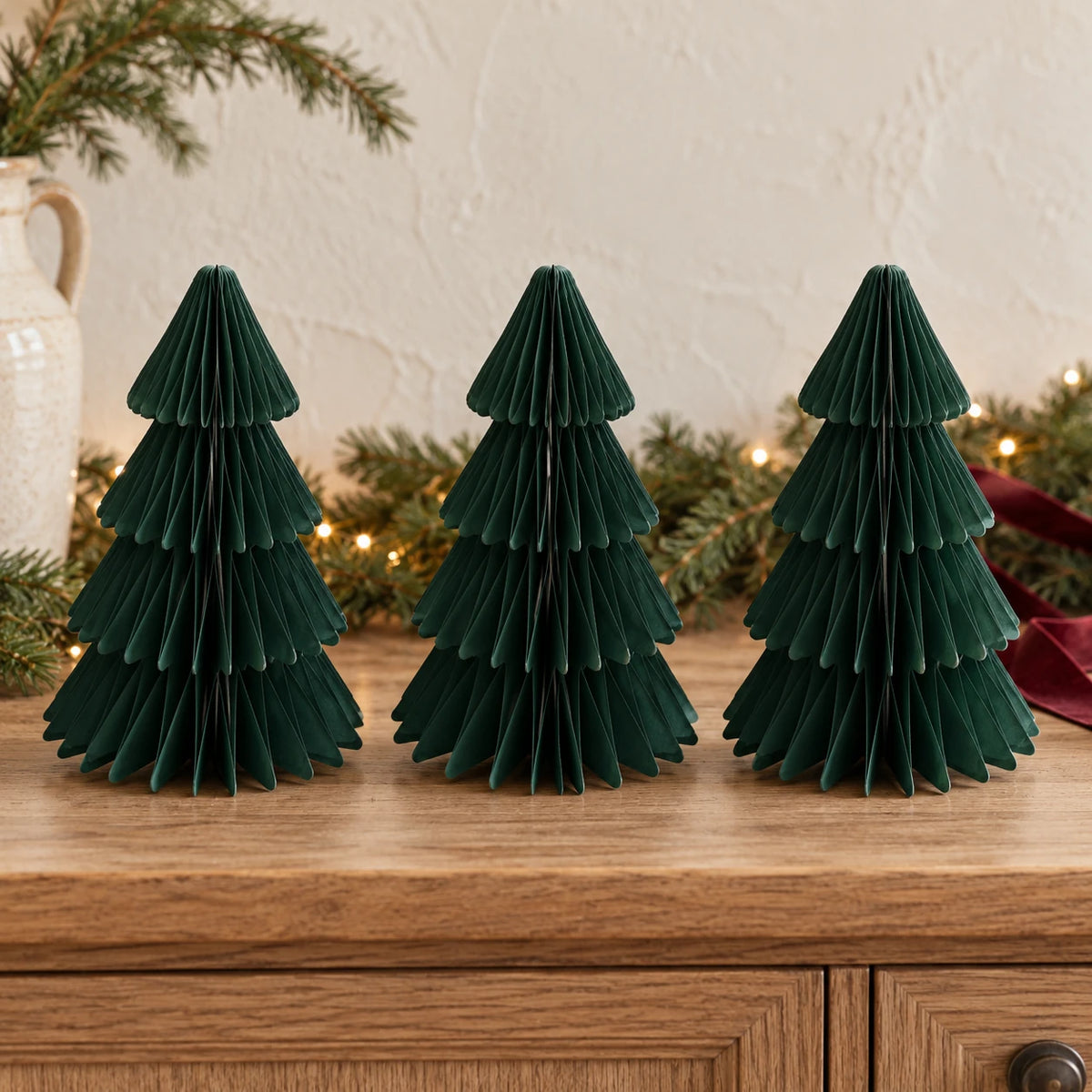 Three matching 20cm dark green paper Christmas trees on an oak sideboard with festive greenery.