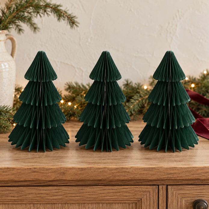 Three matching 20cm dark green paper Christmas trees on an oak sideboard with festive greenery.