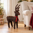 Small standing brown Salim deer with antlers and red ribbon beside an armchair