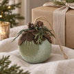 Green velvet woodland bauble on cream linen beside a wrapped gift.
