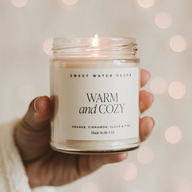 Hand holding a lit Sweet Water Decor Warm and Cozy 9 oz candle in a clear jar against soft festive lights.