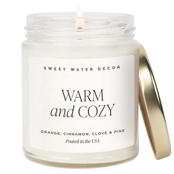 Lit Warm and Cozy 9 oz candle in a clear glass jar with a gold-coloured lid beside it on white.