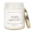 Lit Warm and Cozy 9 oz candle in a clear glass jar with a gold-coloured lid beside it on white.