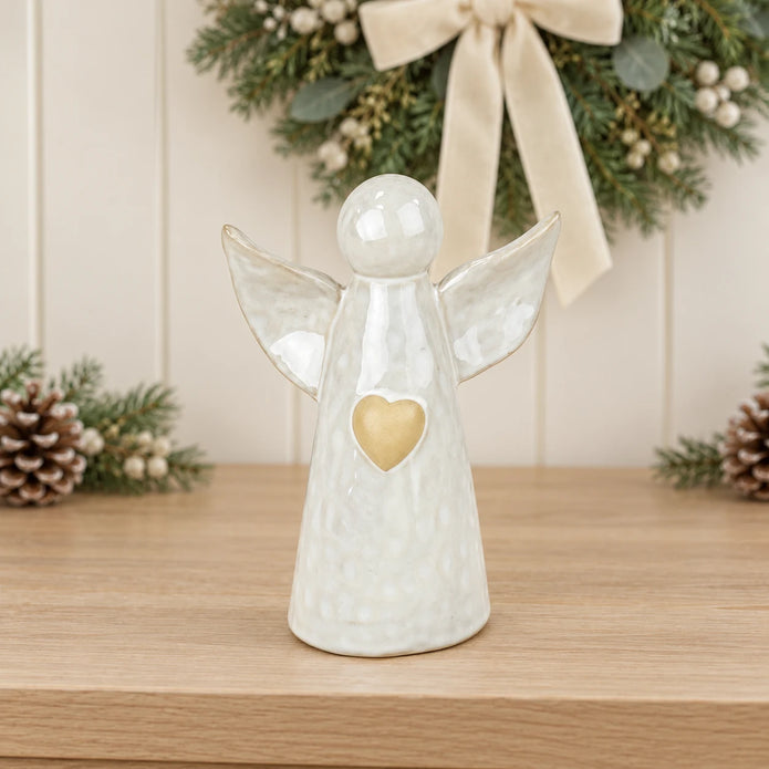 White ceramic angel ornament with pale gold heart styled on a neutral console with Christmas greenery and cream accents.