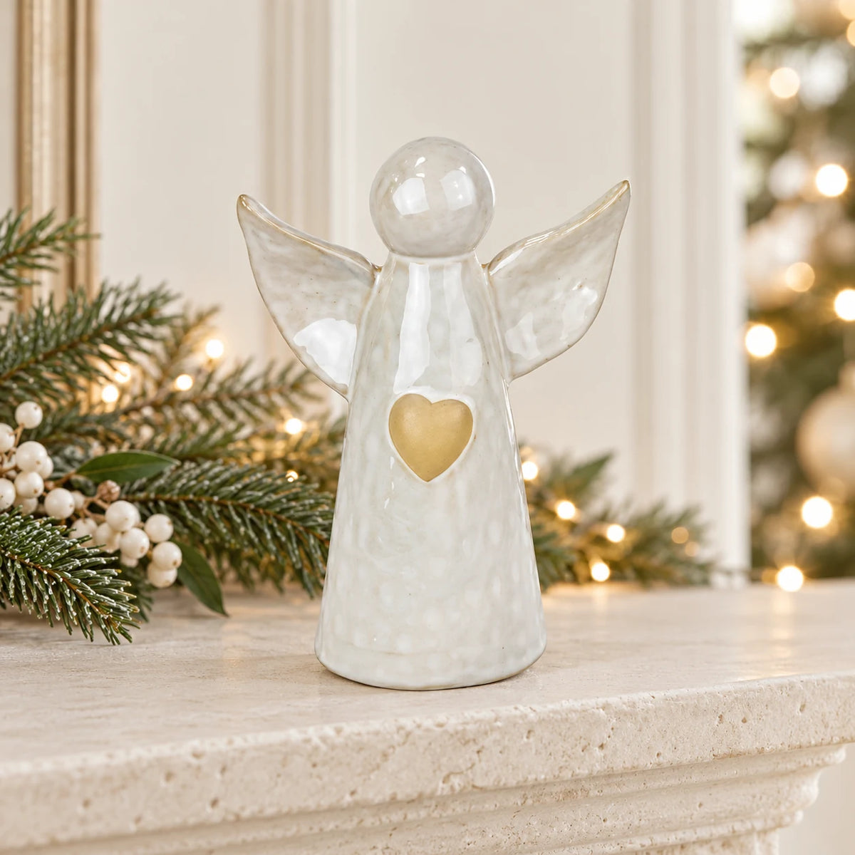 White ceramic angel ornament with pale gold heart styled on a neutral mantel with Christmas greenery and cream accents.