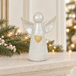 White ceramic angel ornament with pale gold heart styled on a neutral mantel with Christmas greenery and cream accents.
