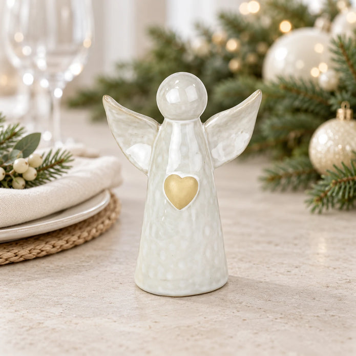 White ceramic angel ornament with pale gold heart styled on a neutral table with Christmas greenery and cream accents.