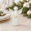 White ceramic angel ornament with pale gold heart styled on a neutral table with Christmas greenery and cream accents.