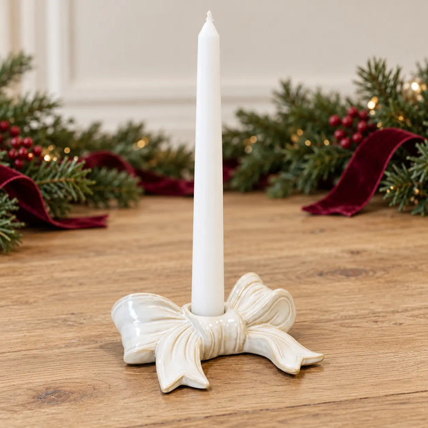 White ceramic bow candle holder with an unlit white taper and Christmas greenery in the background