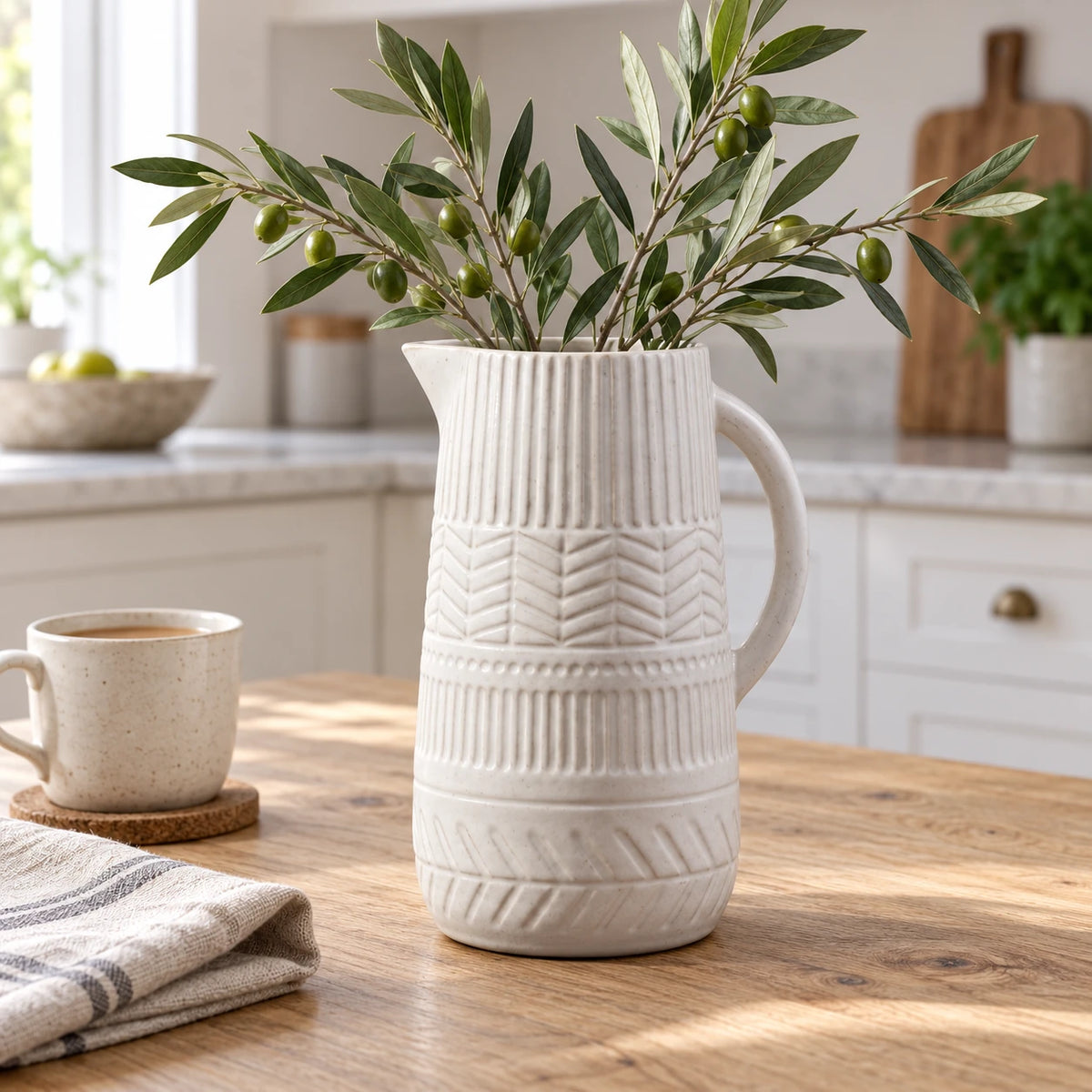 White ceramic decorative jug with olive foliage on a light oak kitchen table.