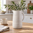 White ceramic decorative jug with olive foliage on a light oak kitchen table.