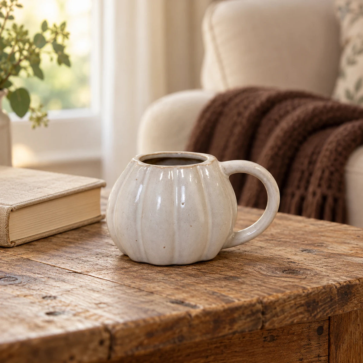 White ceramic pumpkin mug from Satchville, ideal for autumn decor, shown on a living room table with cozy accents.