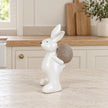 Large white ceramic rabbit holding a taupe egg, 21cm tall, on a pale stone counter in a bright neutral kitchen.