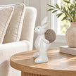 Large white ceramic rabbit holding a taupe egg, 21cm tall, on an oak side table beside a cream linen armchair.