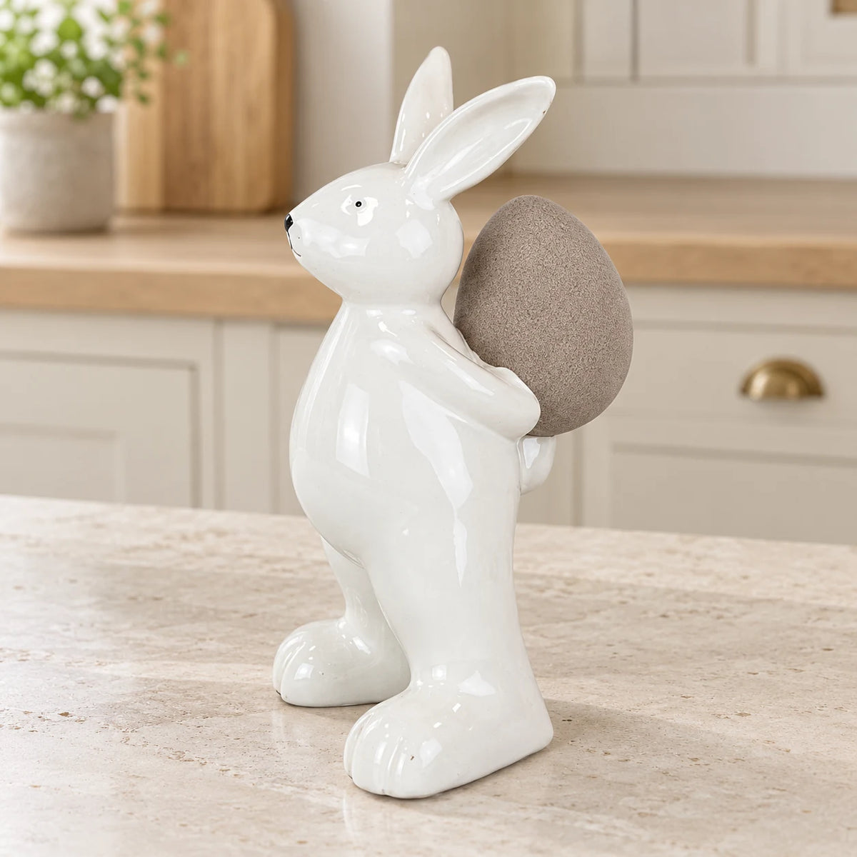 Small white ceramic rabbit holding a taupe egg, 18cm tall, on a pale stone counter in a bright neutral kitchen.