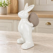 Small white ceramic rabbit holding a taupe egg, 18cm tall, on a pale stone counter in a bright neutral kitchen.