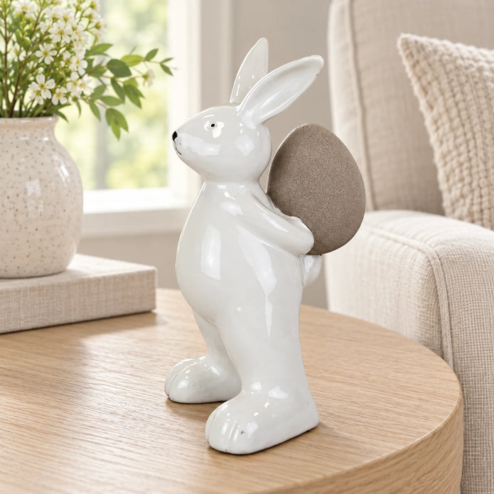 Small white ceramic rabbit holding a taupe egg, 18cm tall, on an oak side table beside a cream linen armchair.
