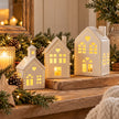 Three white ceramic light-up houses styled with pinecones and warm festive lights