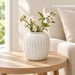 Small warm white ceramic leaf vase styled with white blossom on a pale oak side table beside a cream linen armchair.