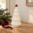 White ceramic LED tree with red star glowing on an oak console with fir and burgundy ribbon.