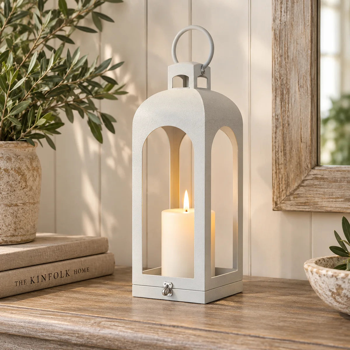 White metal open-faced lantern with LED pillar candle styled on wooden console alongside olive foliage