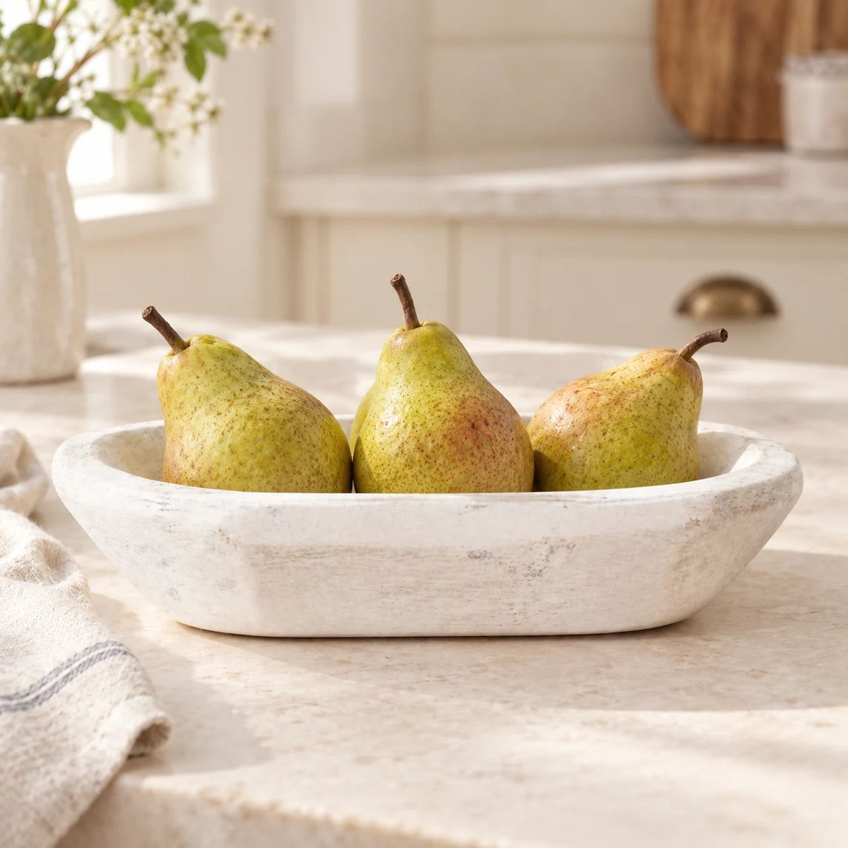 White mini dough bowl holding three pears on a cream kitchen worktop, with folded linen beside it.