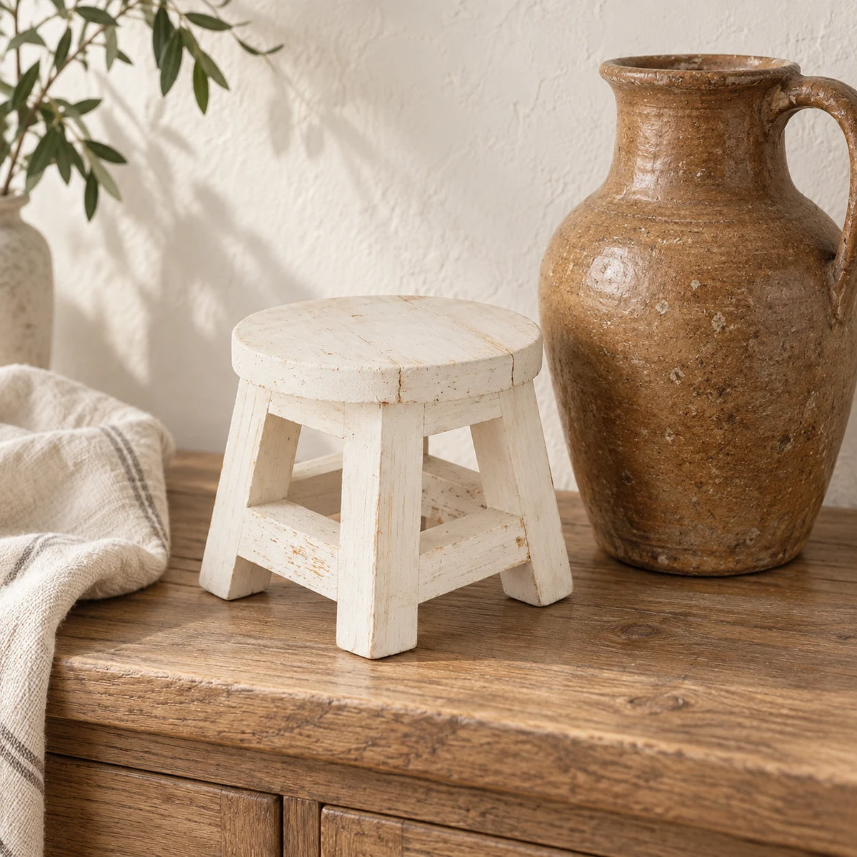 White wooden mini stool styled empty beside a tall brown ceramic jug on an oak console.
