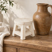 White wooden mini stool styled empty beside a tall brown ceramic jug on an oak console.
