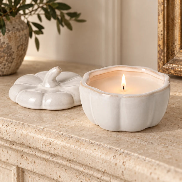 White porcelain pumpkin candle burning on a cream mantel with its lid beside it.