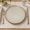 Overhead view of white stoneware dinner plate on ivory linen with cutlery and water glass