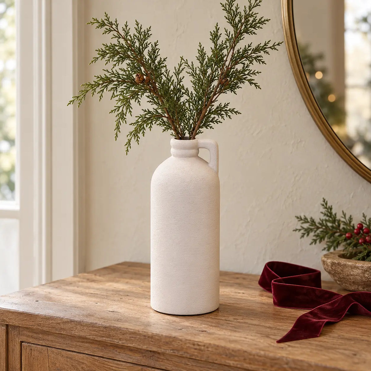 White textured bottle vase with small handle and faux cedar stems on a Christmas console