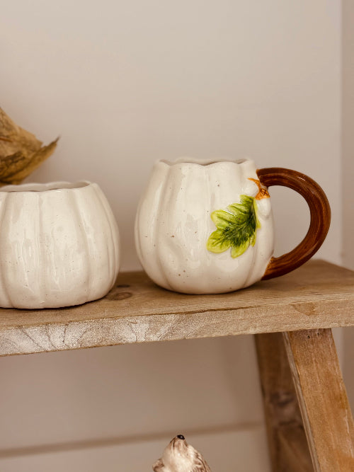 Two ceramic pumpkin mugs on a wooden shelf with a neutral background