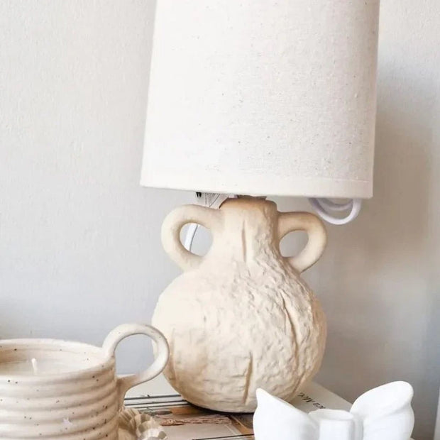 Small cream ceramic table lamp with a ceramic handle and linen shade, perfect for cozy lighting in living spaces.