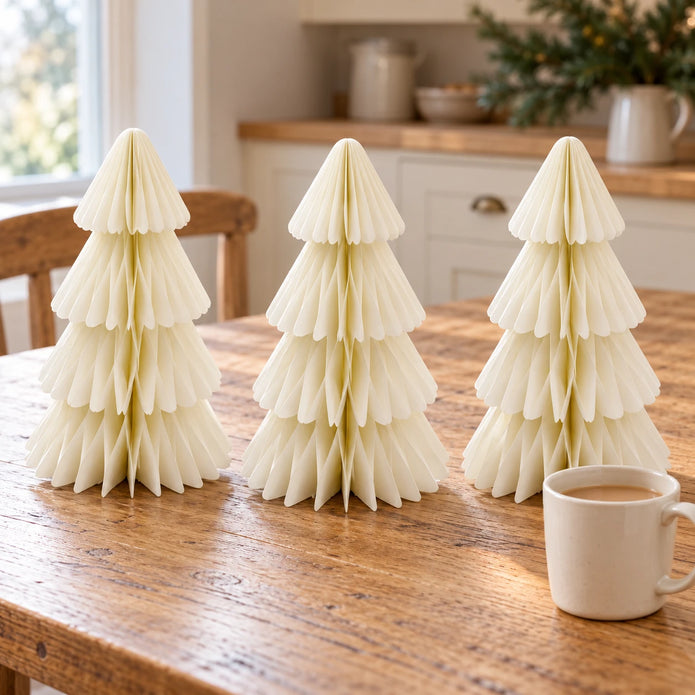 Set of three winter white paper Christmas trees on an oak kitchen table beside a mug of tea.
