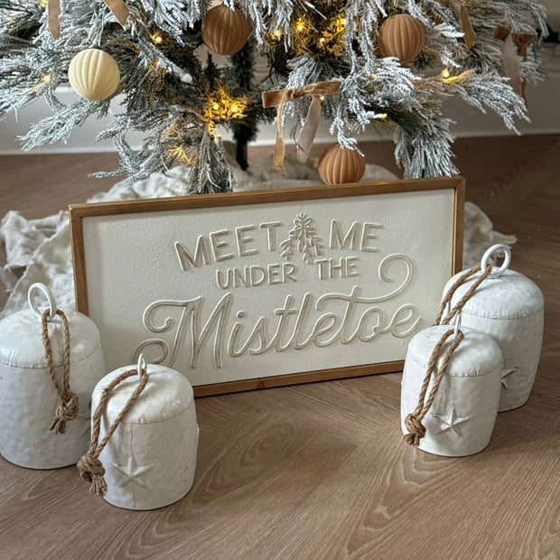 White Meet Me Under the Mistletoe framed sign beside a Christmas tree and bells